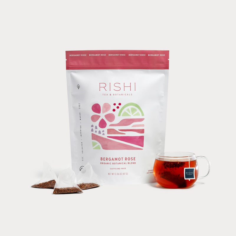 A bag of Rishi Tea & Botanicals Bergamot Rose tea is shown with three pyramid sachets and a clear glass cup of aromatic bergamot tea blended with rose petals, all set on a white background.