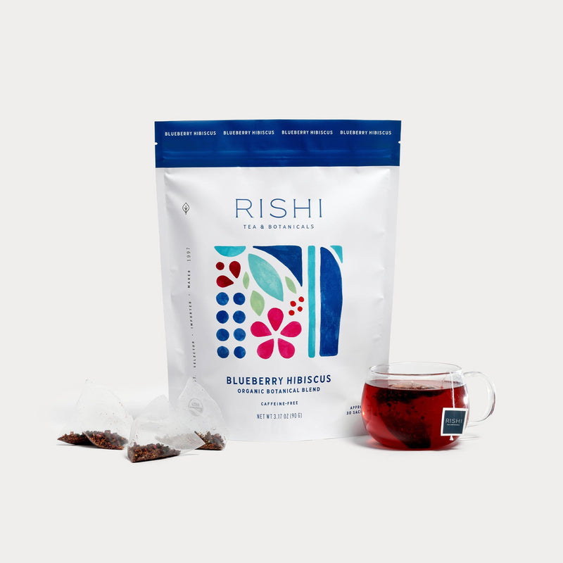 A package of Rishi Tea & Botanicals Blueberry Hibiscus tea is shown with a clear mug of brewed hibiscus tea and two sachets, one opened, all set against a white background.
