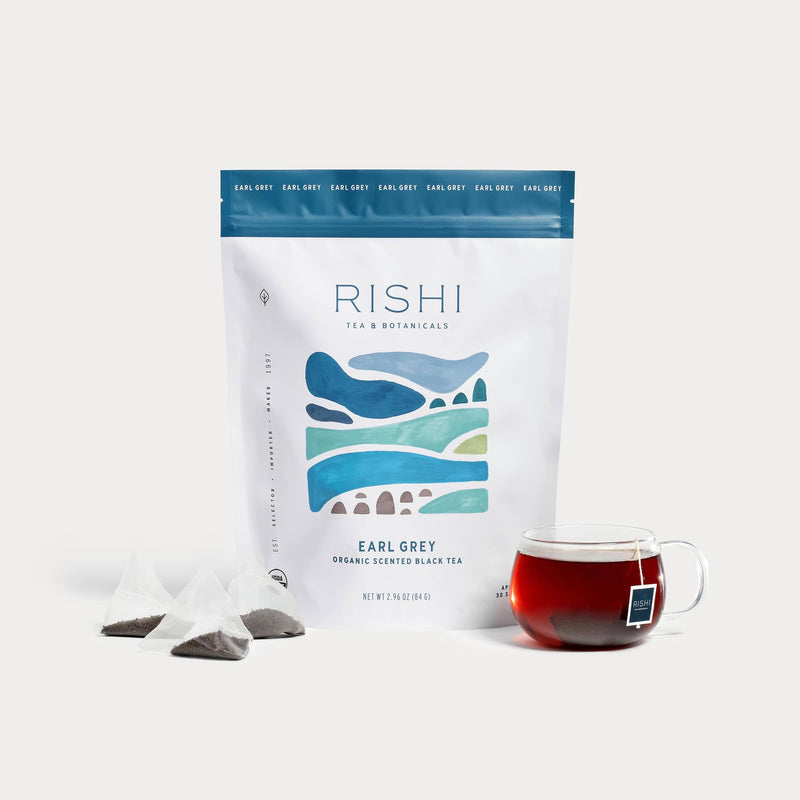 A bag of Rishi Tea & Botanicals Organic Earl Grey Black Tea, infused with bergamot oil, stands behind three pyramid sachets and a glass cup of brewed tea with a tea bag and tag.