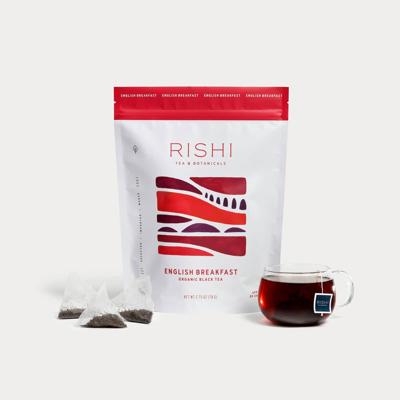 A bag of Rishi Tea & Botanicals English Breakfast organic black tea, three tea bags, and a clear glass cup filled with malty, chocolatey tea are displayed against a white background.