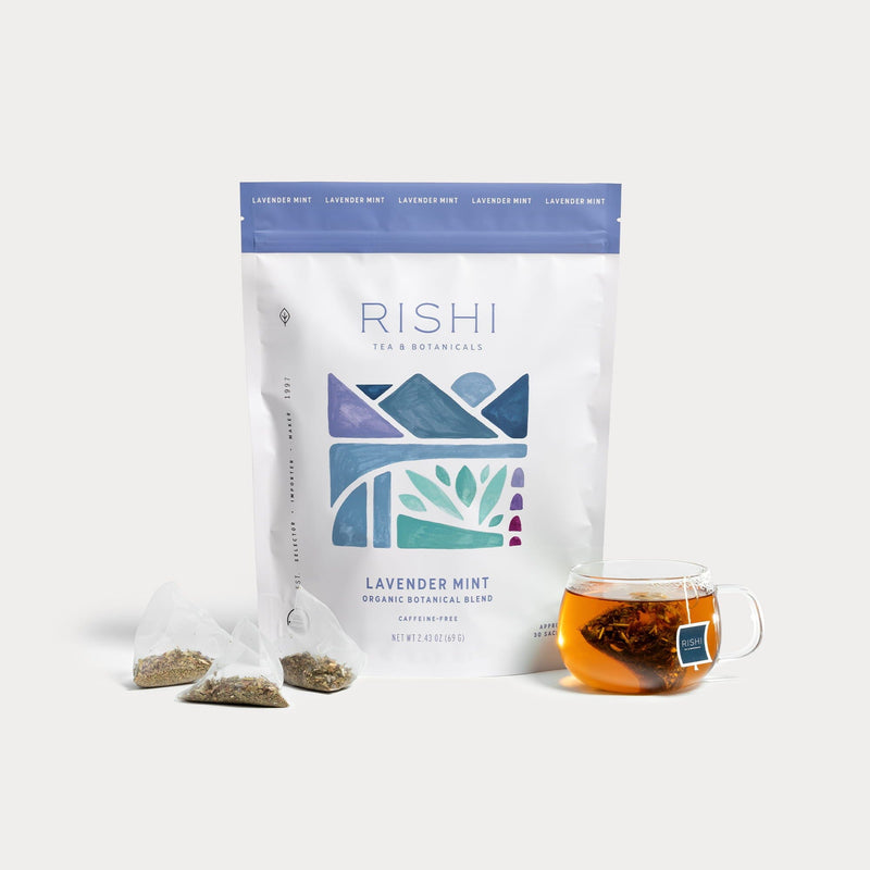 A bag of Rishi Tea & Botanicals Lavender Mint with three pyramid tea bags and a glass cup of calming lavender and peppermint-infused tea on a white background.