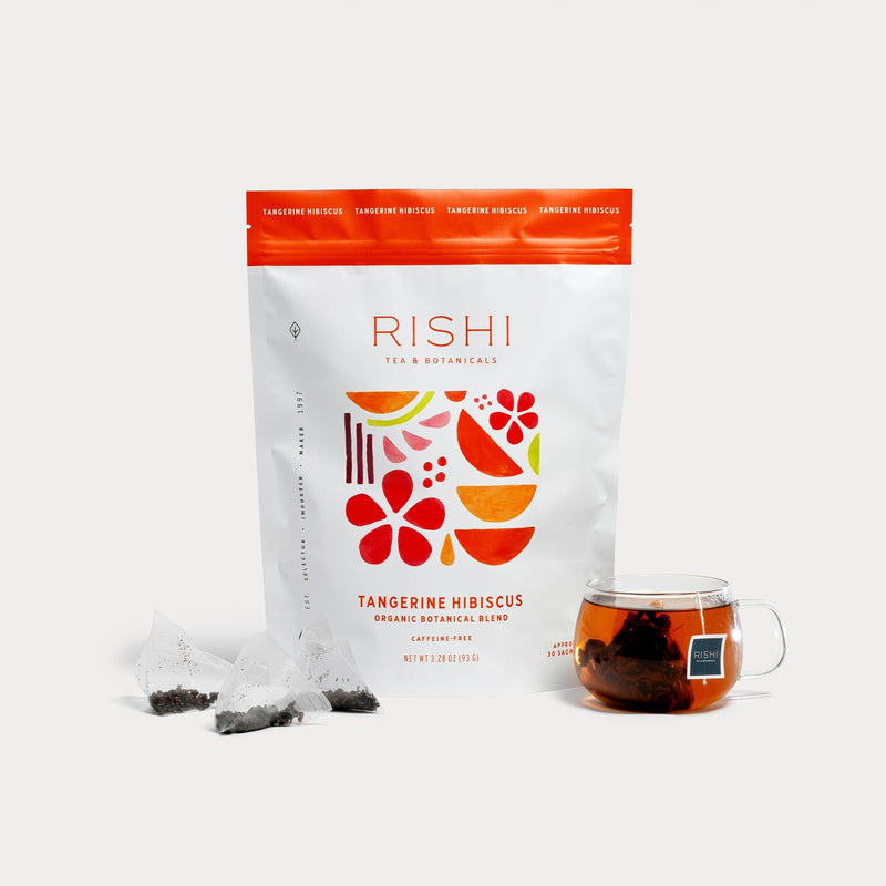 A bag of Rishi Tea & Botanicals Tangerine Hibiscus stands beside a glass of brewed hibiscus tea and two pyramid tea bags on a white background—an inviting, caffeine-free blend ideal for any time of day.