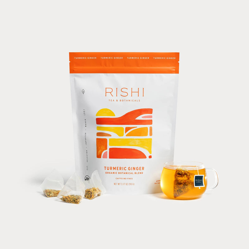 A package of Rishi Tea & Botanicals Turmeric Ginger Ayurvedic herbal tea is displayed with three pyramid tea bags and a glass cup of brewed tea in front of the box.