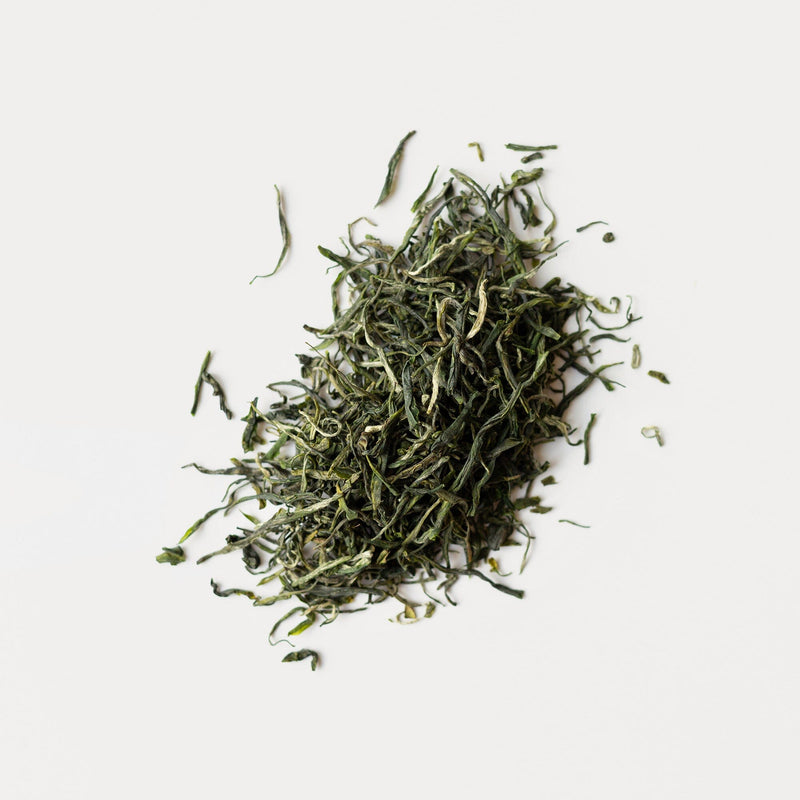 A pile of loose Green Needles Qun Ti Zhong tea leaves scattered on a white background. (Rishi Tea & Botanicals)