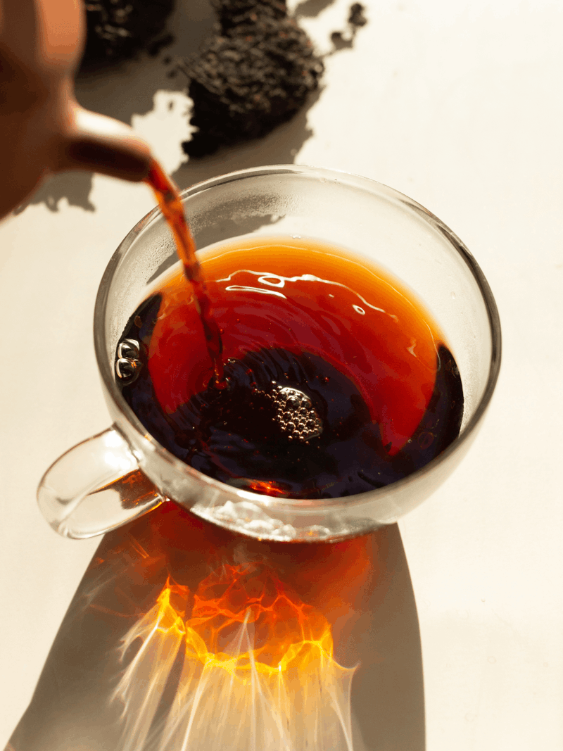 A glass cup filled with chamomile tea, with several chamomile flowers floating on top and a few lying on the wet surface nearby.