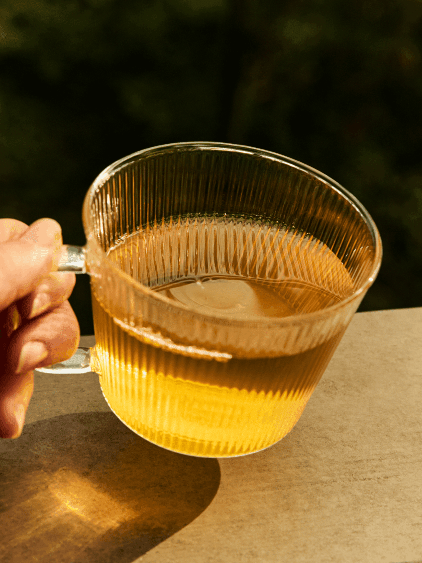 A glass cup filled with light brown tea is placed on a beige surface next to a gold spoon holding loose tea leaves.